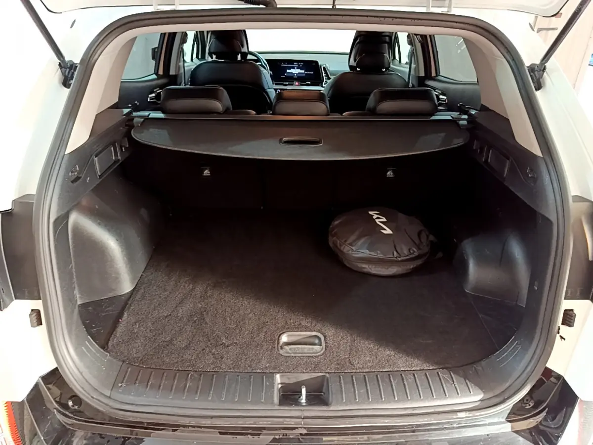 Coffre ouvert d'un KIA SPORTAGE blanc vu de l'arrière, avec cache-bagages et sac noir marqué KIA visible à l'intérieur.