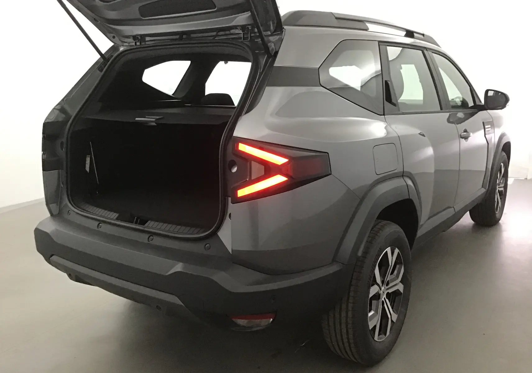 Vue 3/4 arrière droit du Dacia Bigster gris schiste avec coffre ouvert et feux arrière allumés dans un studio.