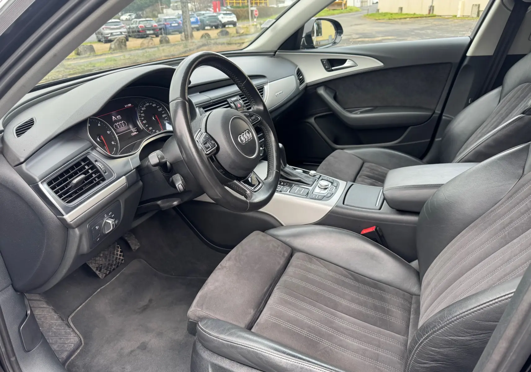 Vue intérieure côté conducteur de l'Audi A6 Avant 2015 noire, avec sièges cuir et alcantara, volant multifonction et tableau de bord digital.