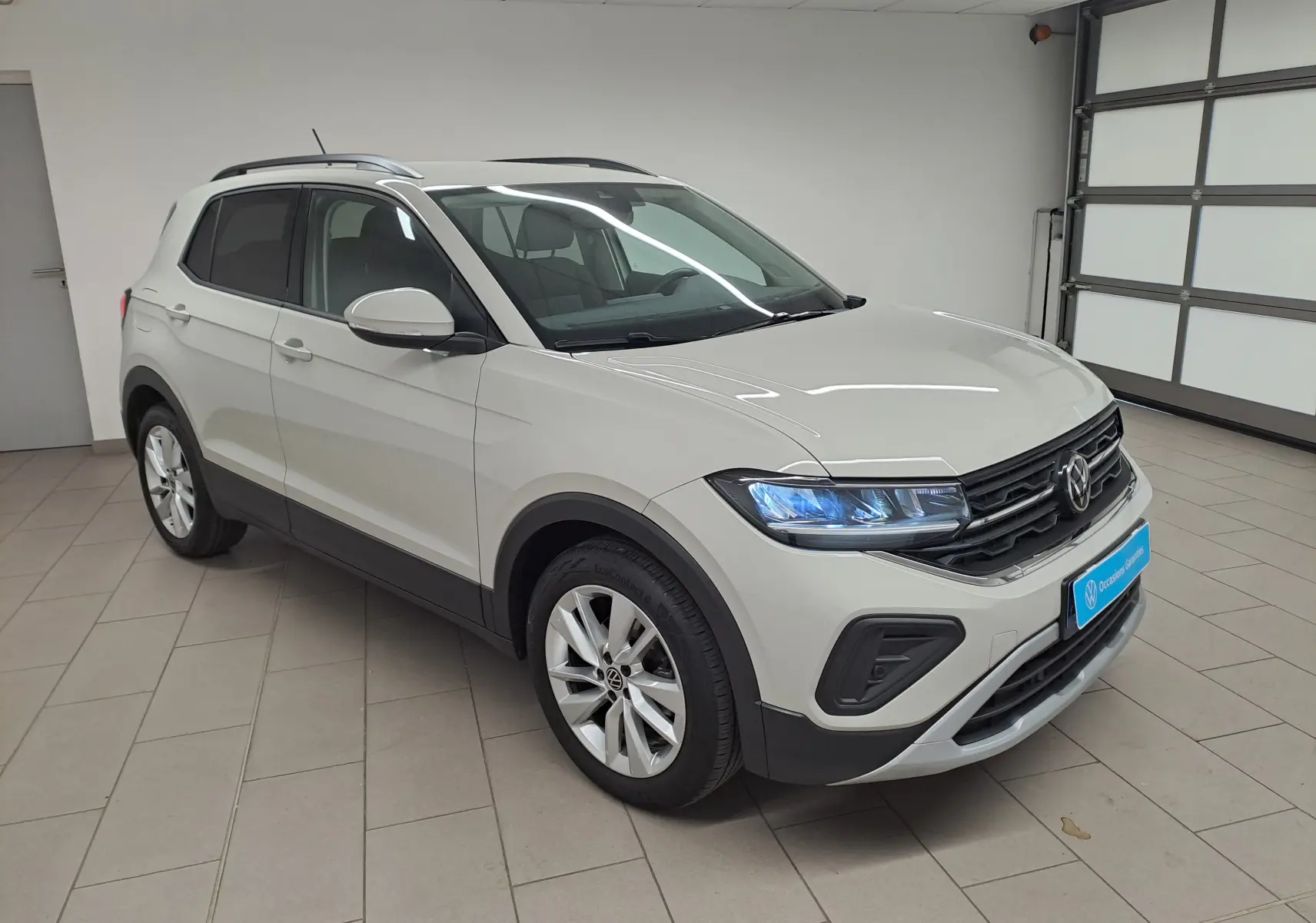 Volkswagen T-Cross Gris Ascot en 3/4 avant droit, avec phares allumés et jantes alliage visibles en intérieur fermé.