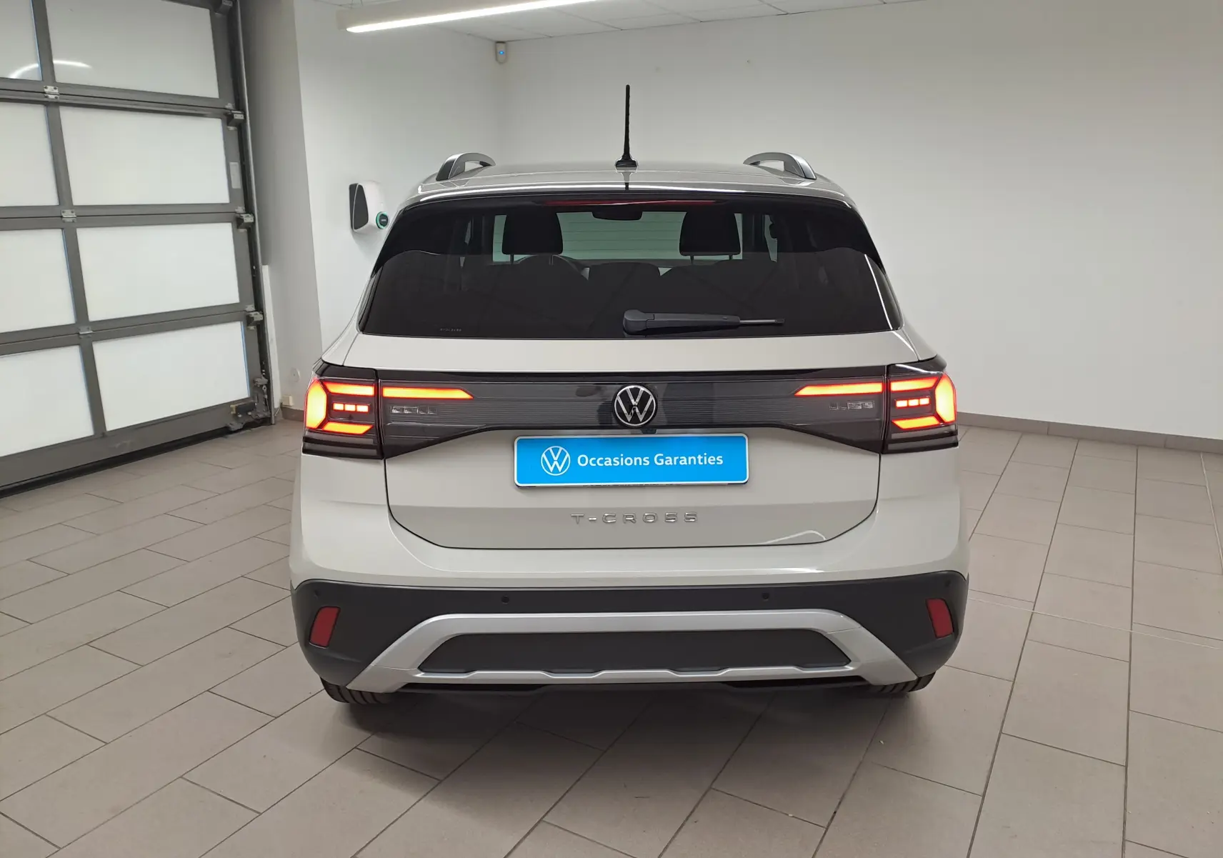 Vue arrière d'un Volkswagen T-Cross 2024 gris Ascot avec feux allumés et logo VW visible en intérieur showroom.