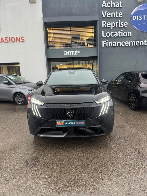 Vue de face du Peugeot 3008 Hybrid noir avec feux diurnes LED en forme de griffes, garé devant un bâtiment commercial.