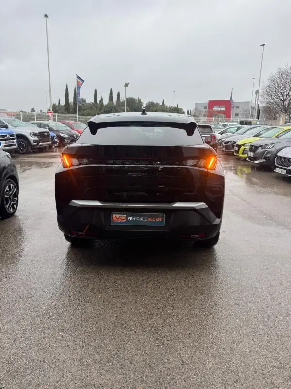 Vue arrière d'un Peugeot 3008 Hybrid 145 noir avec feux allumés, garé sur un parking humide entouré d'autres véhicules.