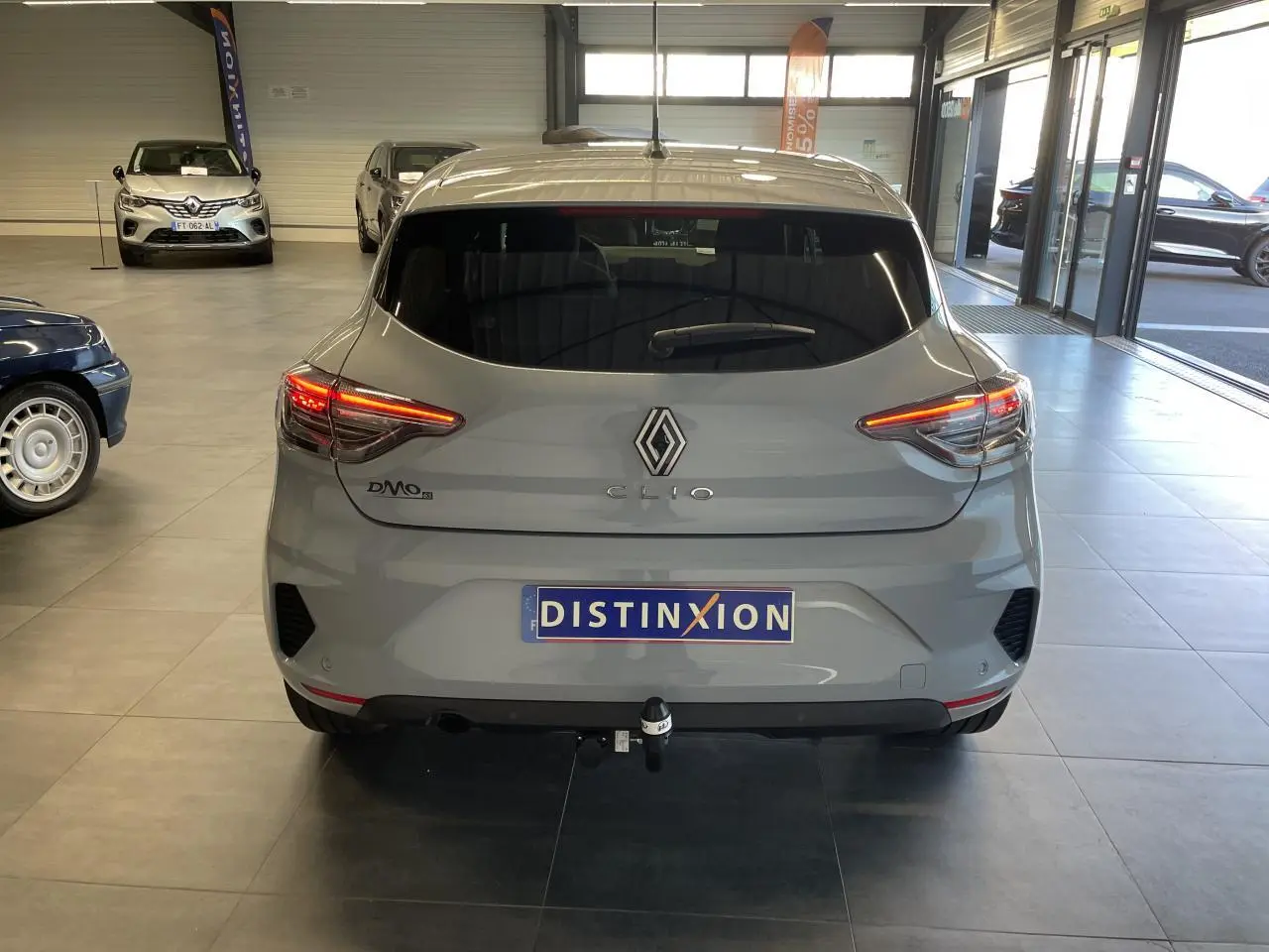Vue arrière d'une Renault Clio gris Rafale 2025 avec feux LED allumés et attelage visible en intérieur showroom.