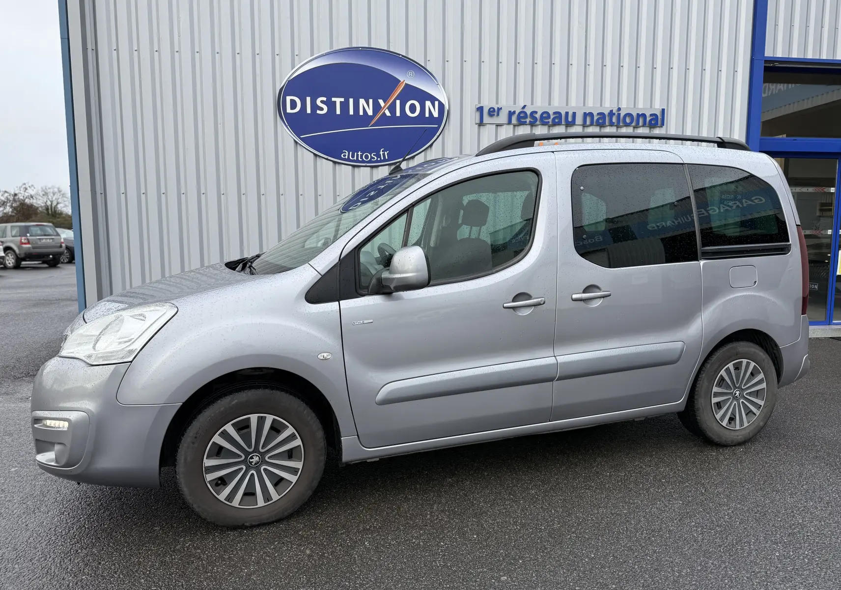 Vue de profil côté gauche d'un Peugeot Partner Tepee gris 2018 garé devant un bâtiment avec logo Distinxion.