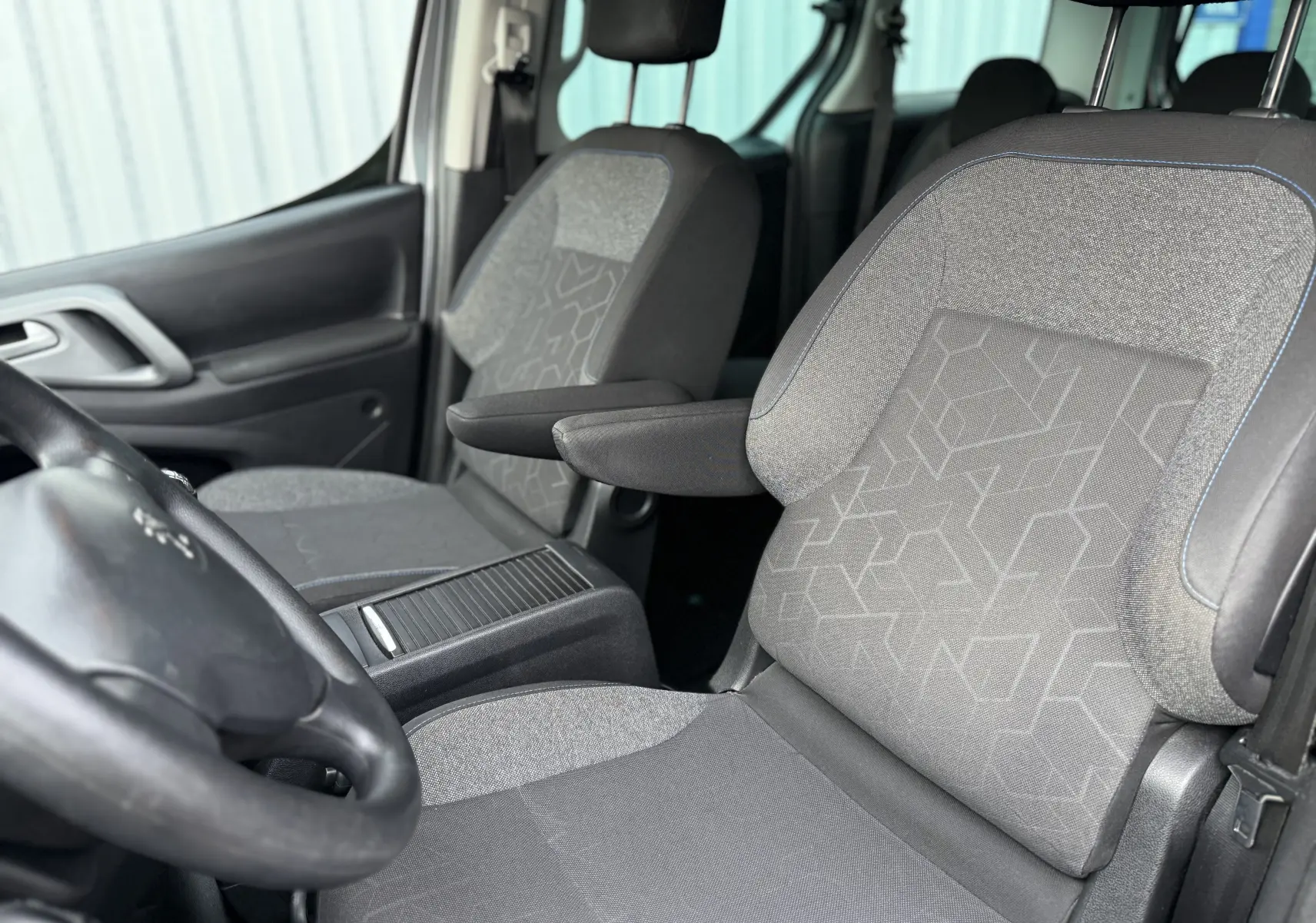 Intérieur du Peugeot Partner Tepee 2018, vue rapprochée des sièges avant en tissu gris avec accoudoirs et volant visible.