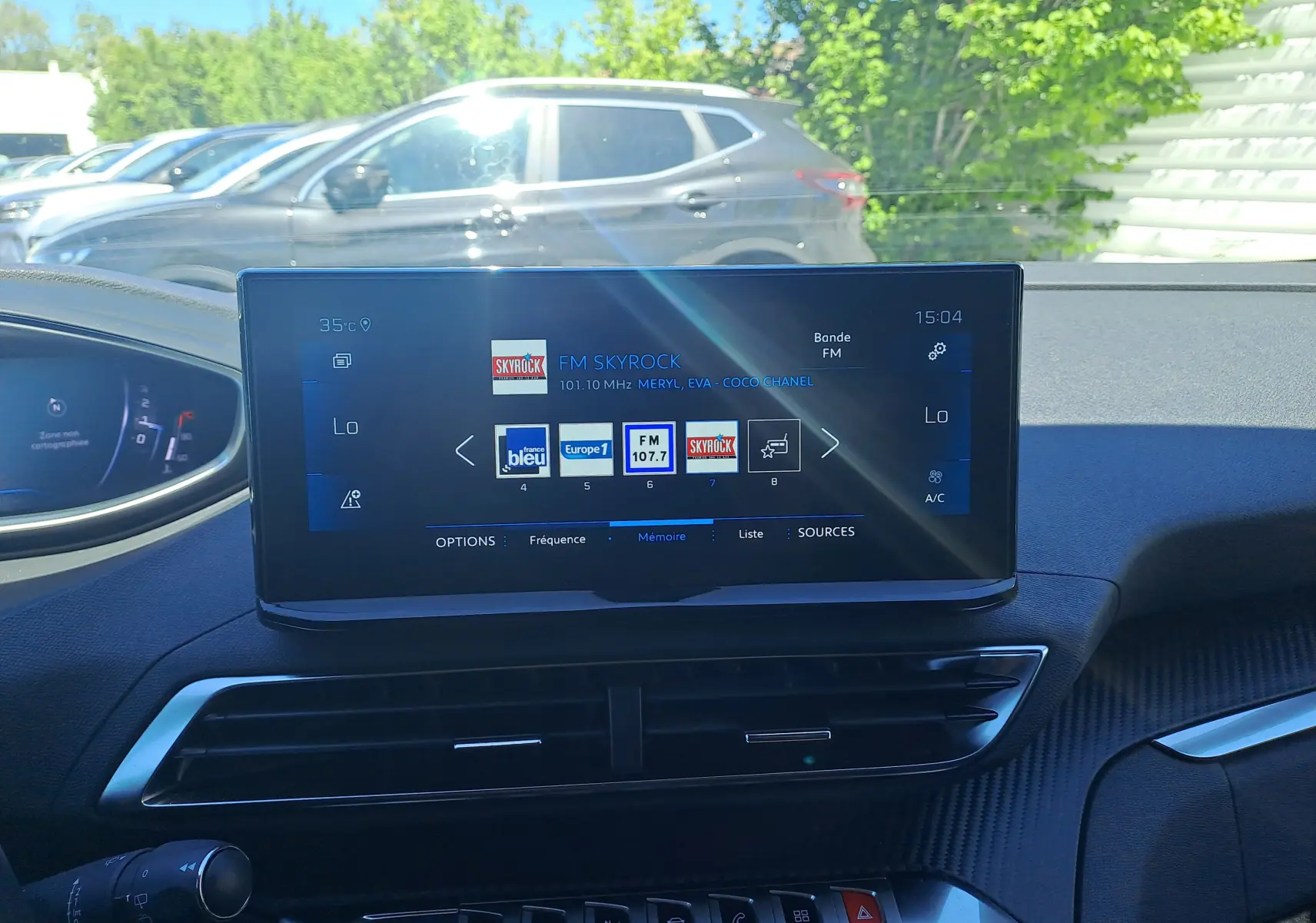 Vue intérieure du tableau de bord du Peugeot 3008 2023, écran central tactile affichant les radios FM, avec extérieur et voiture grise en arrière-plan.
