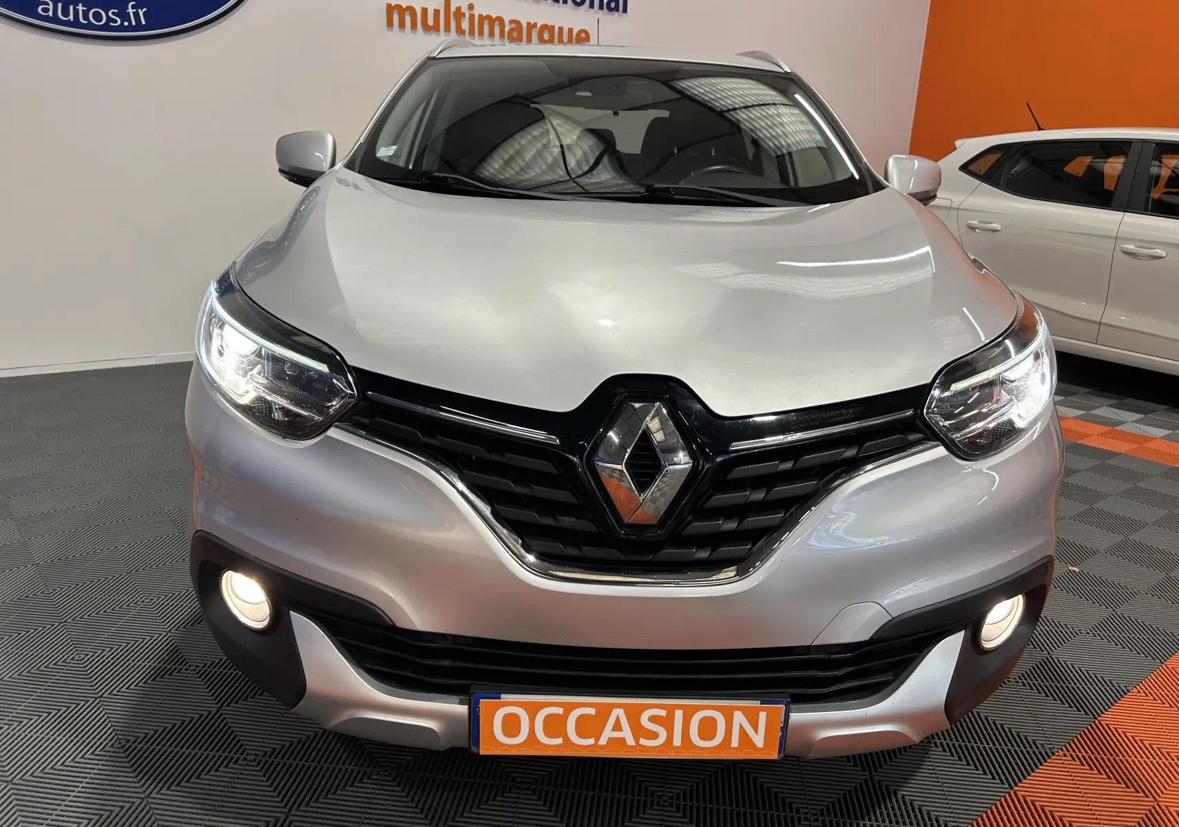 Vue avant d'un Renault Kadjar gris avec phares allumés et plaque "OCCASION" en intérieur showroom.