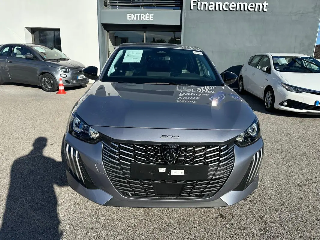 Vue avant d'une Peugeot 208 Hybrid 110 gris métallisé avec calandre noire et phares LED, stationnée devant un bâtiment.