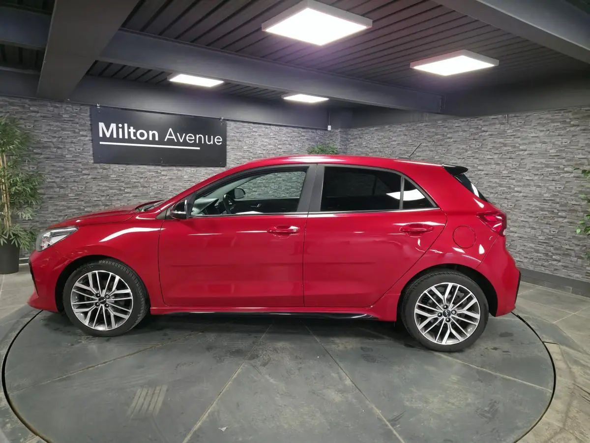 Profil droit d'une KIA RIO rouge métallisée 2022 avec jantes alliage 17'' et vitres surteintées en intérieur showroom.