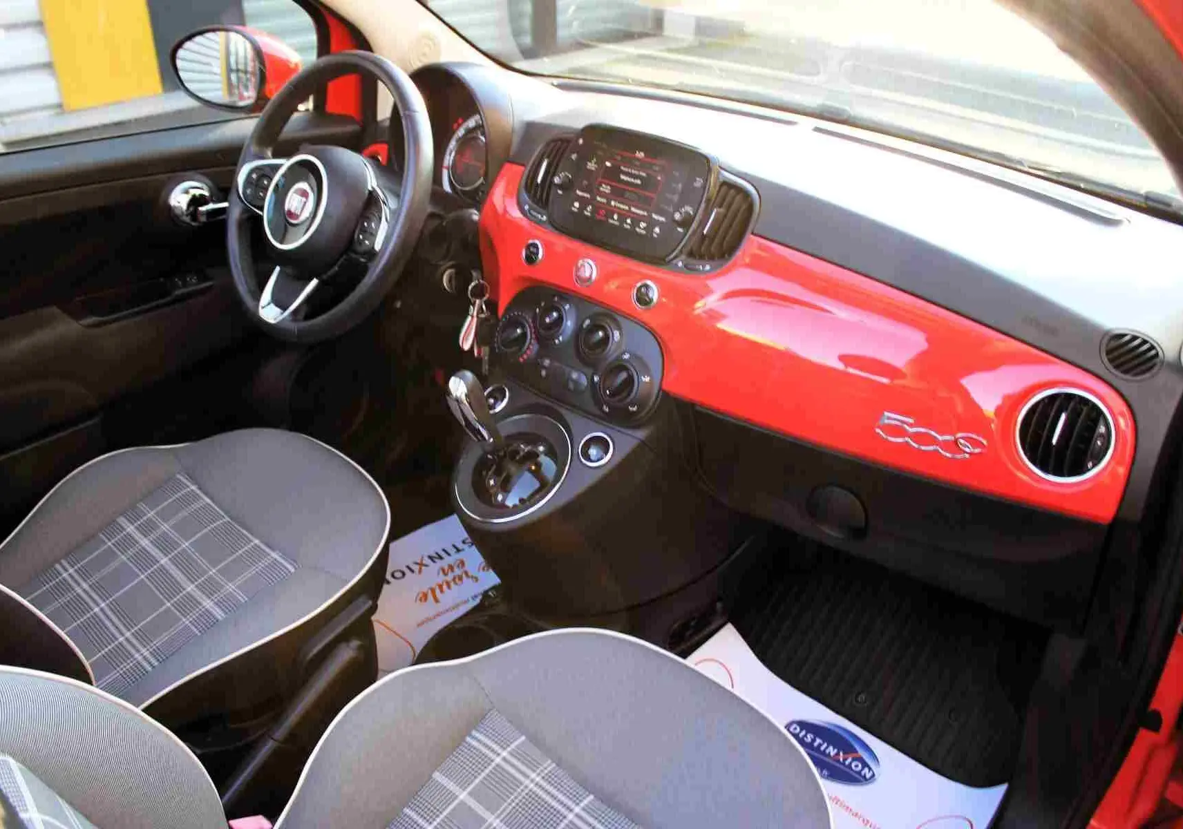 Vue intérieure avant centrée sur le volant et le tableau de bord rouge pastel d'une Fiat 500C 2018 avec écran tactile.