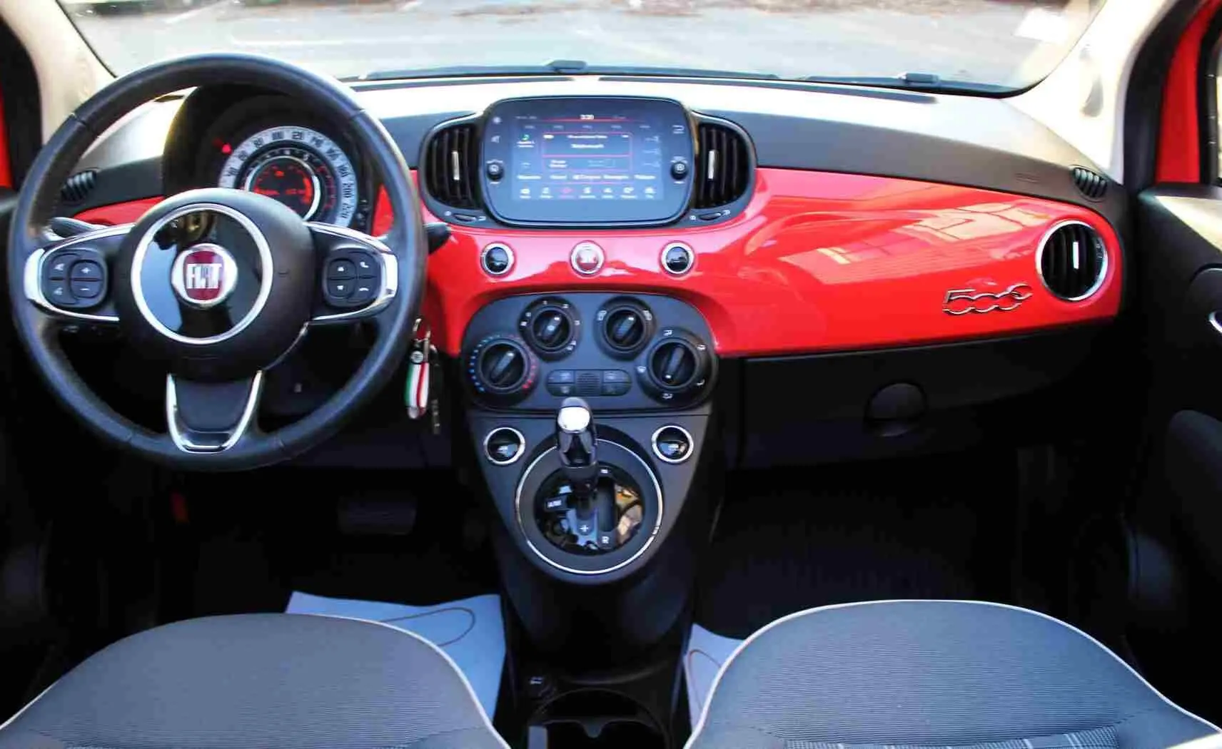 Vue intérieure avant du tableau de bord rouge pastel coral de la Fiat 500C 2018 avec volant multifonction et écran tactile central.