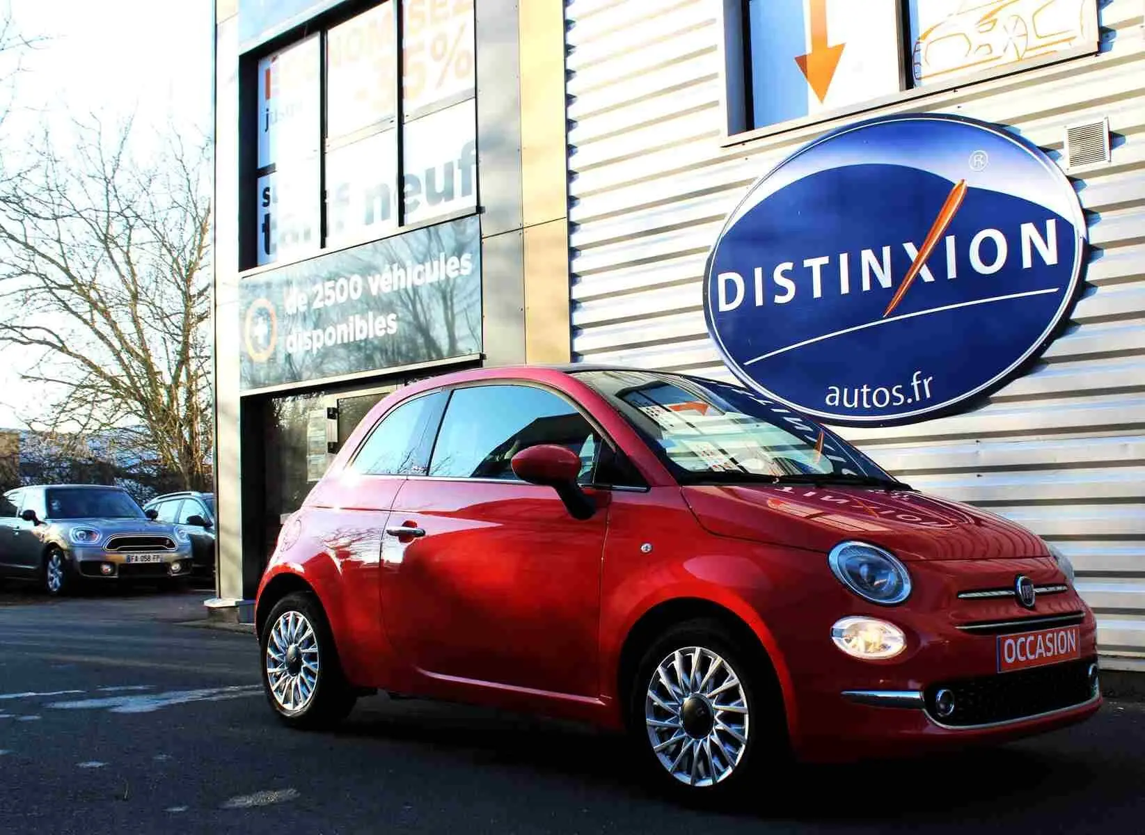 Vue 3/4 avant droite d'une FIAT 500C 2018 rouge pastel avec phares allumés et plaque "OCCASION" devant un concessionnaire.