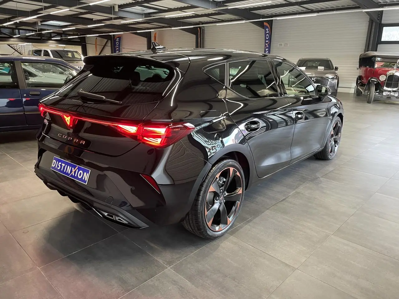 Vue 3/4 arrière droite d'une CUPRA Leon noire avec jantes cuivrées et feux arrière LED allumés en intérieur showroom.