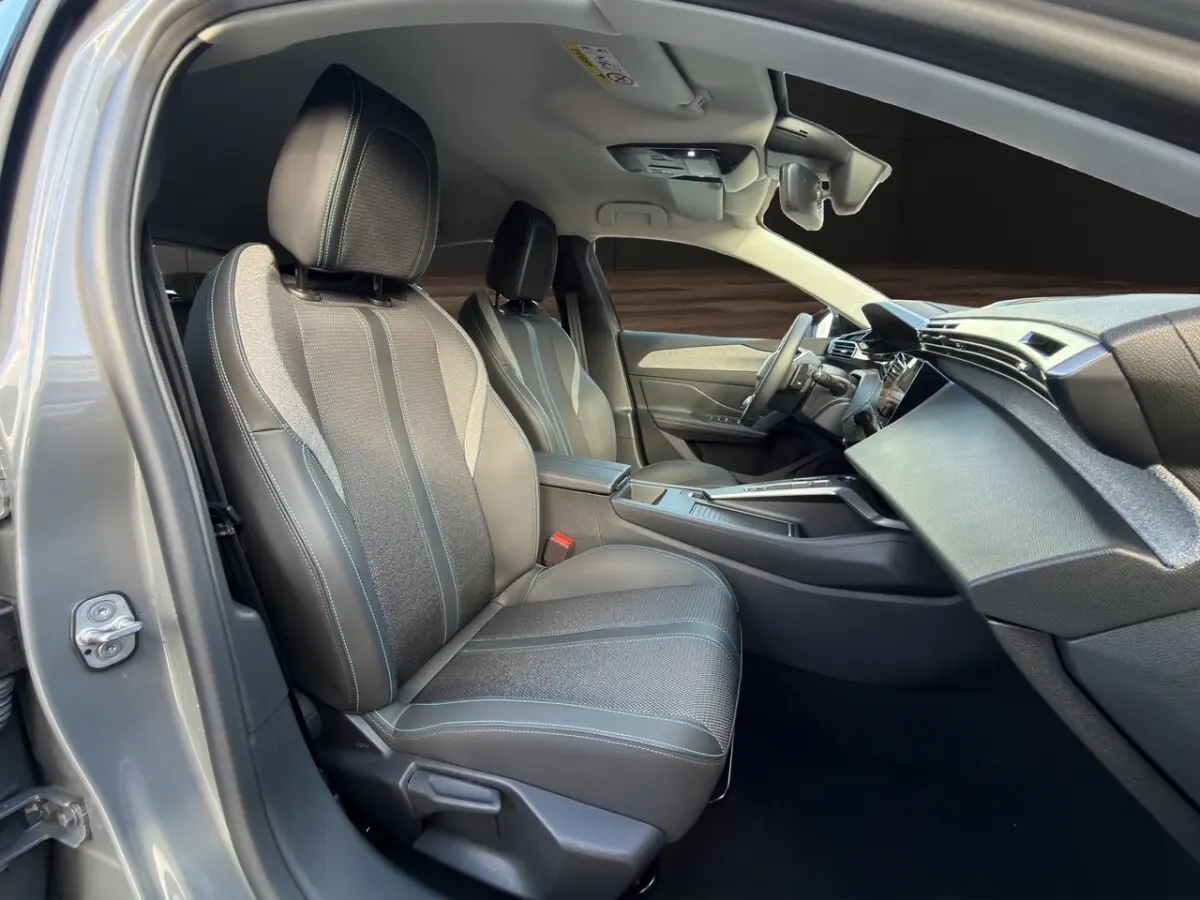 Vue intérieure côté conducteur de la Peugeot 308 Hybrid 145 Allure 2025 gris foncé, sièges en tissu et cuir avec surpiqûres bleues.