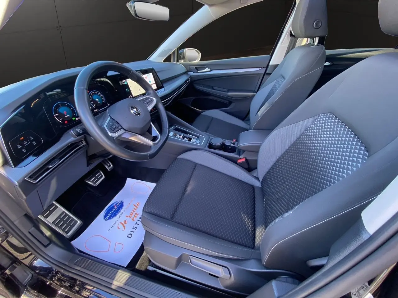 Vue intérieure côté conducteur de la Volkswagen Golf VIII noire, avec tableau de bord numérique et sièges tissu noir.