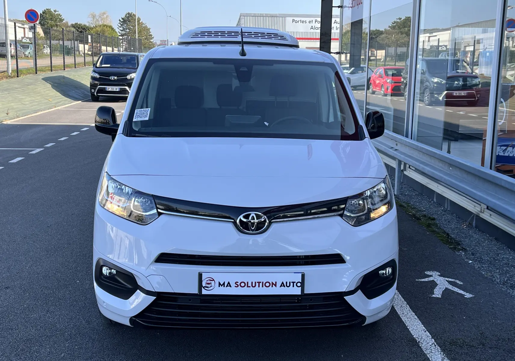 Vue frontale d'un Toyota Proace City blanc glacier avec toit frigorifique et phares allumés, stationné devant un showroom.