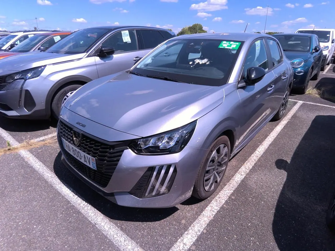 Peugeot 208 gris clair vue 3/4 avant droit, calandre noire et feux LED à signature verticale, stationnée en extérieur.