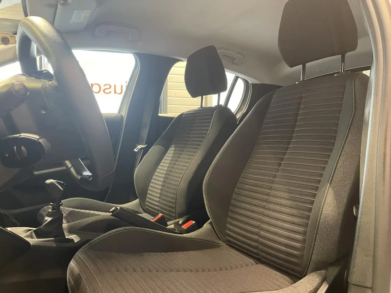 Vue intérieure côté conducteur montrant les sièges avant en tissu noir avec surpiqûres et levier de vitesses manuel.