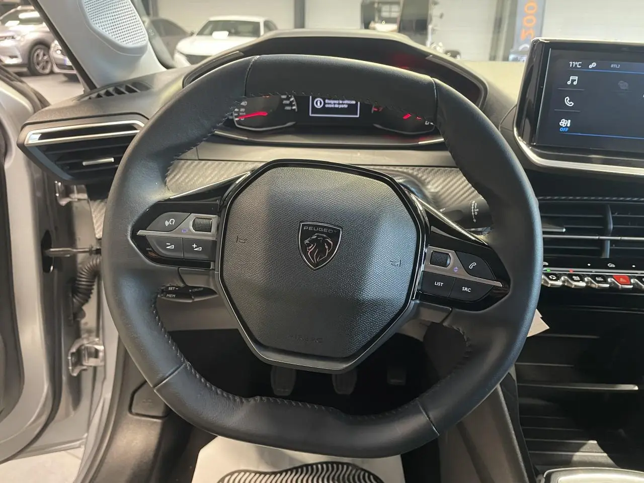 Vue intérieure centrée sur le volant cuir noir de la Peugeot 208 gris clair avec tableau de bord et écran tactile.