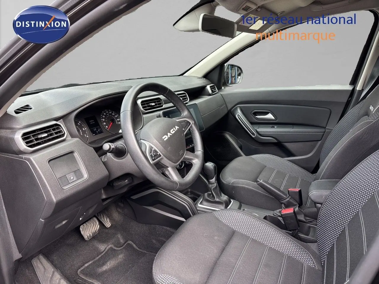 Vue intérieure côté conducteur du Dacia Duster 2024 avec tableau de bord noir et sièges tissu gris foncé.