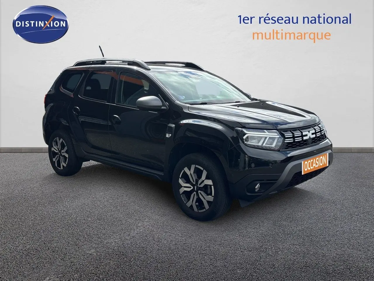 Dacia Duster noir en 3/4 avant droit, avec jantes bi-ton et barres de toit visibles sur fond neutre.