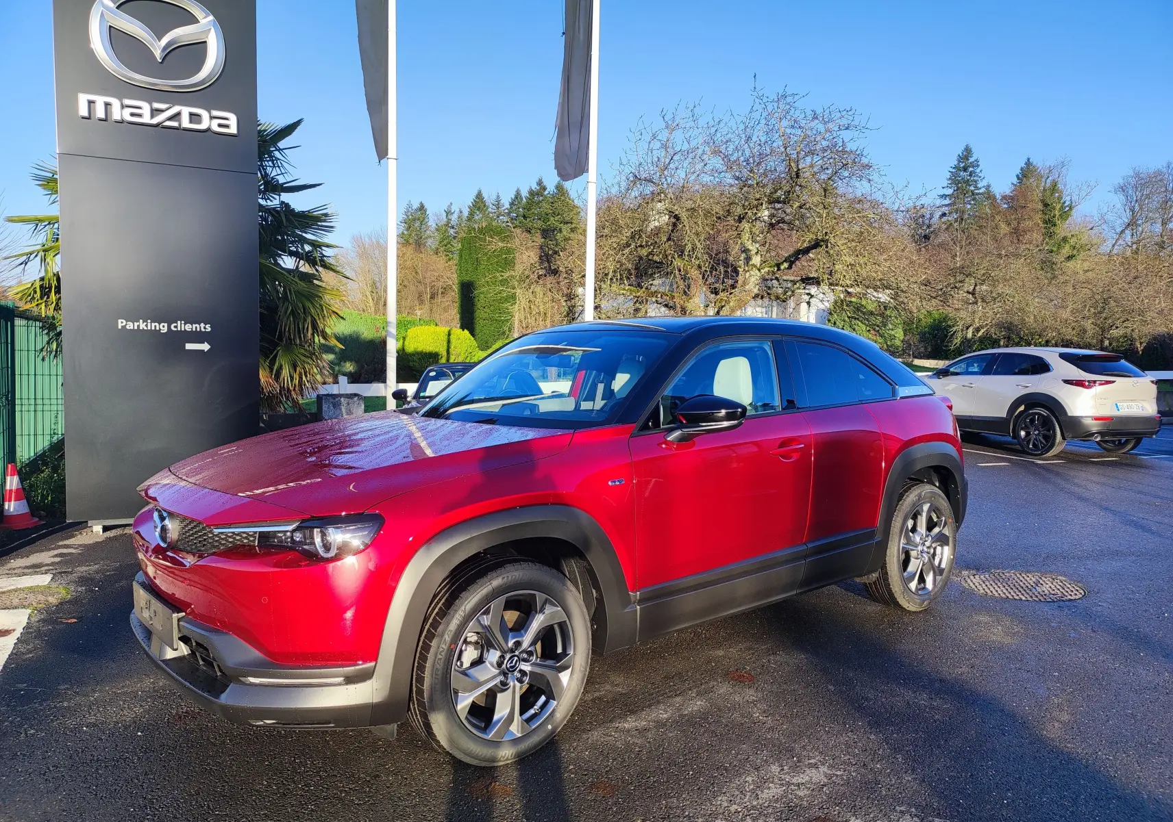 Vue 3/4 avant droite d'un Mazda MX-30 rouge métal avec toit noir, garé devant un panneau Mazda sous un ciel bleu.
