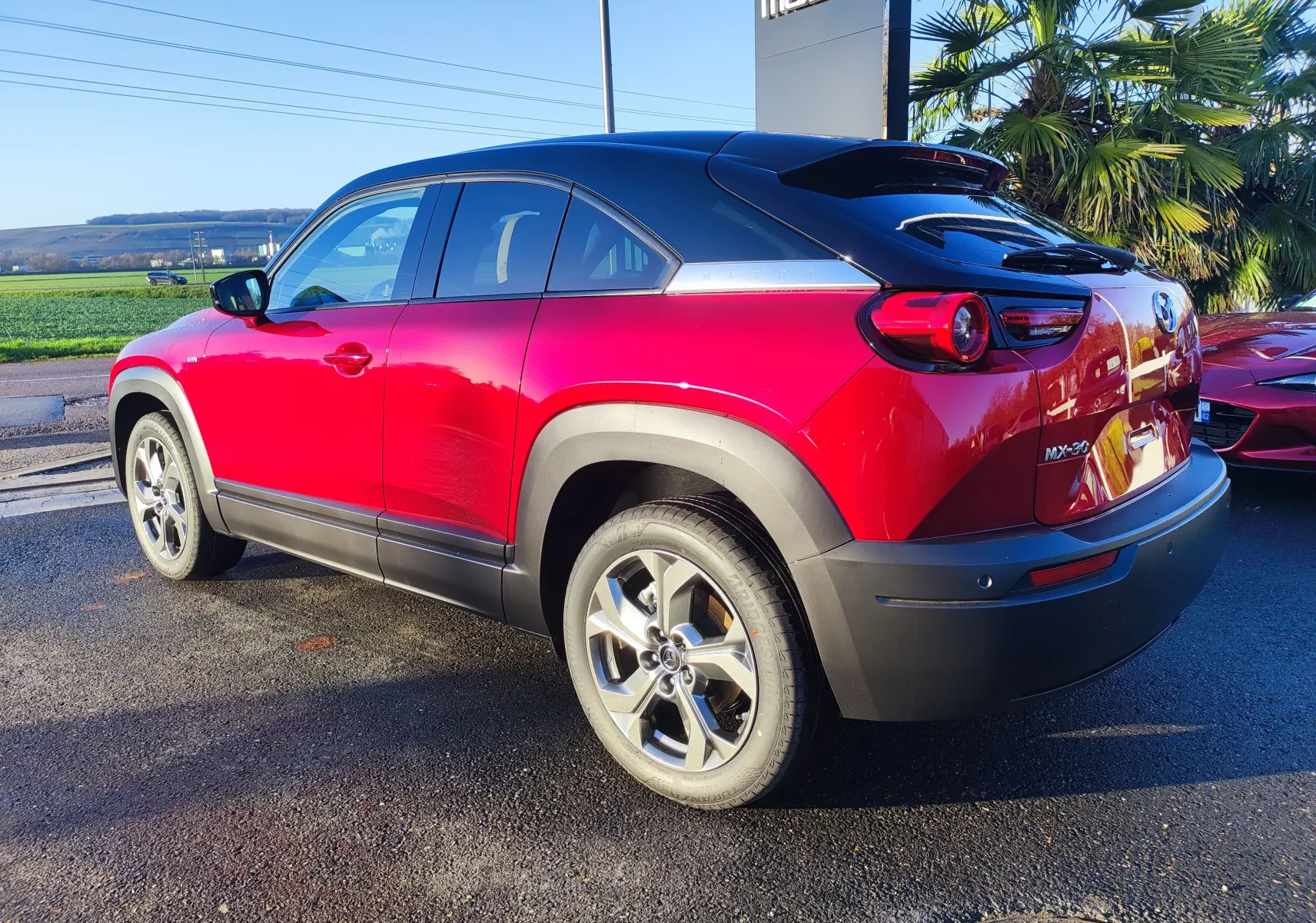 Vue 3/4 arrière droite d'un Mazda MX-30 rouge métal 2023 avec détails noirs et jantes alliage au soleil.