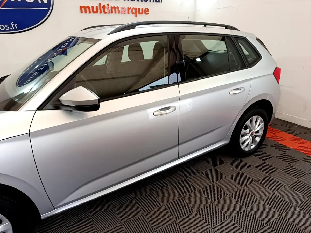 Vue latérale côté gauche d'un Skoda Kamiq gris clair garé en intérieur sur sol à motifs noirs et rouges.