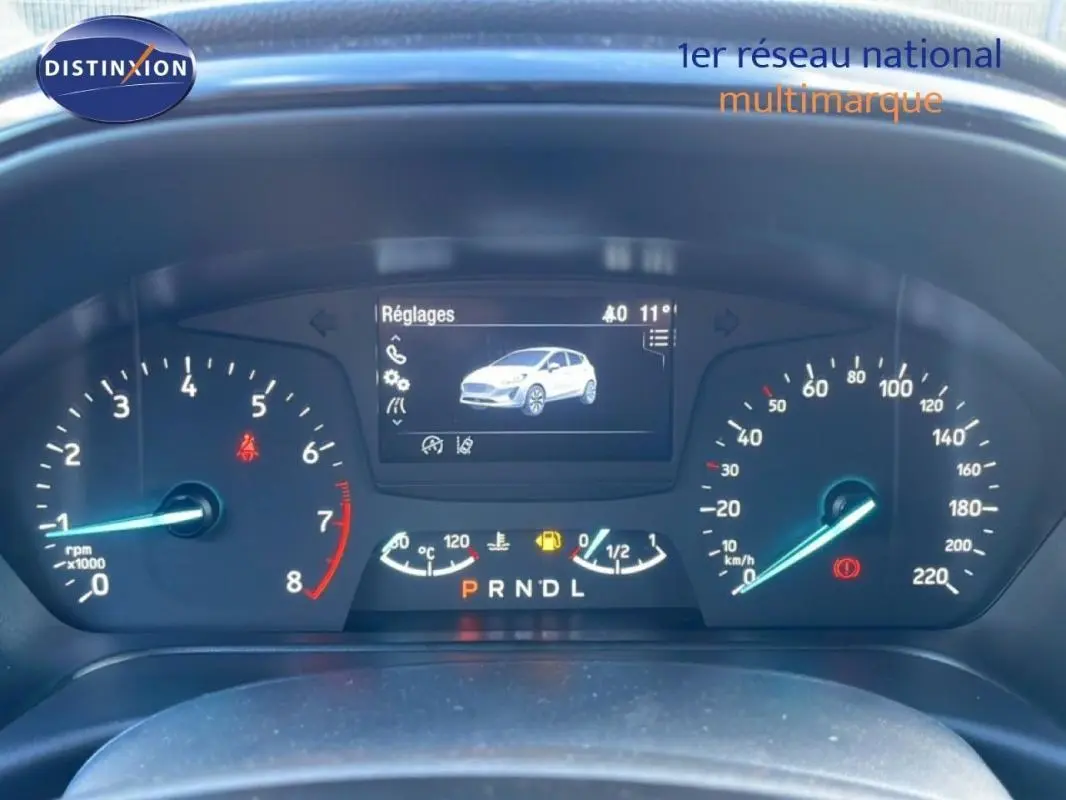 Vue rapprochée du tableau de bord numérique de la Ford Fiesta blanche, affichant les réglages et compteurs.