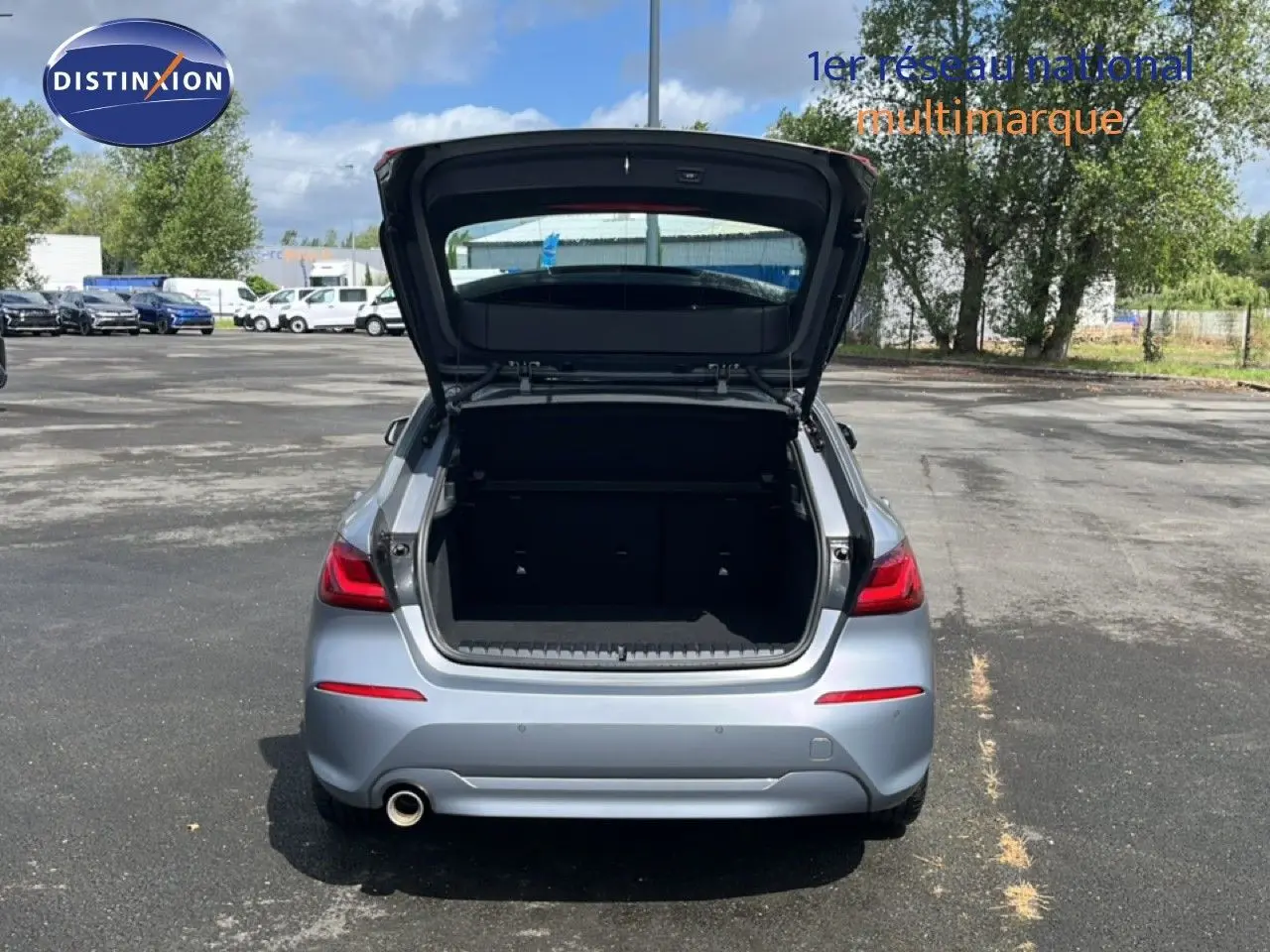 Vue arrière d'une BMW Série 1 116i 2022 gris Frozen Pure avec coffre ouvert sur un parking extérieur.