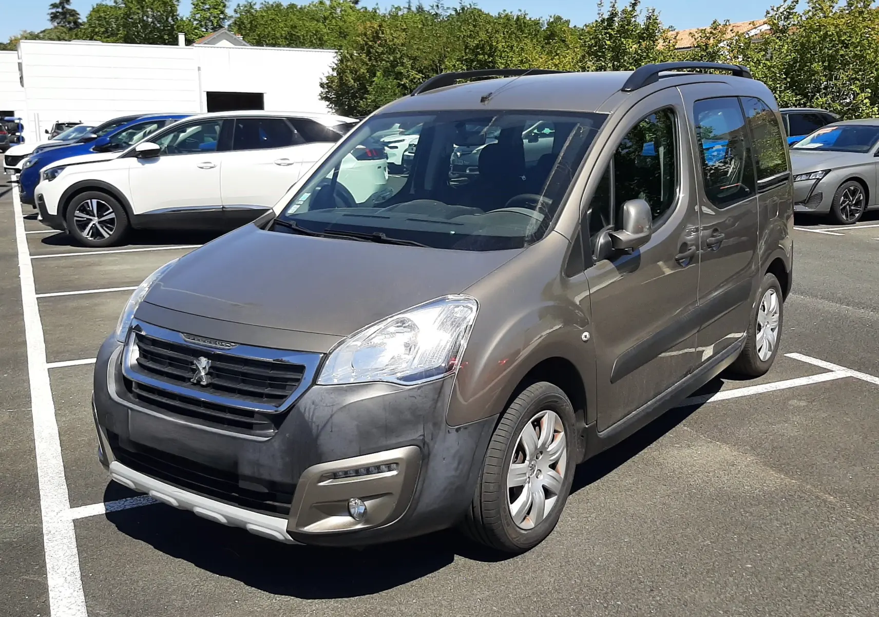 Peugeot Partner Tepee marron vue 3/4 avant droit, avec barres de toit et pare-chocs avant noir mat, stationné en extérieur.