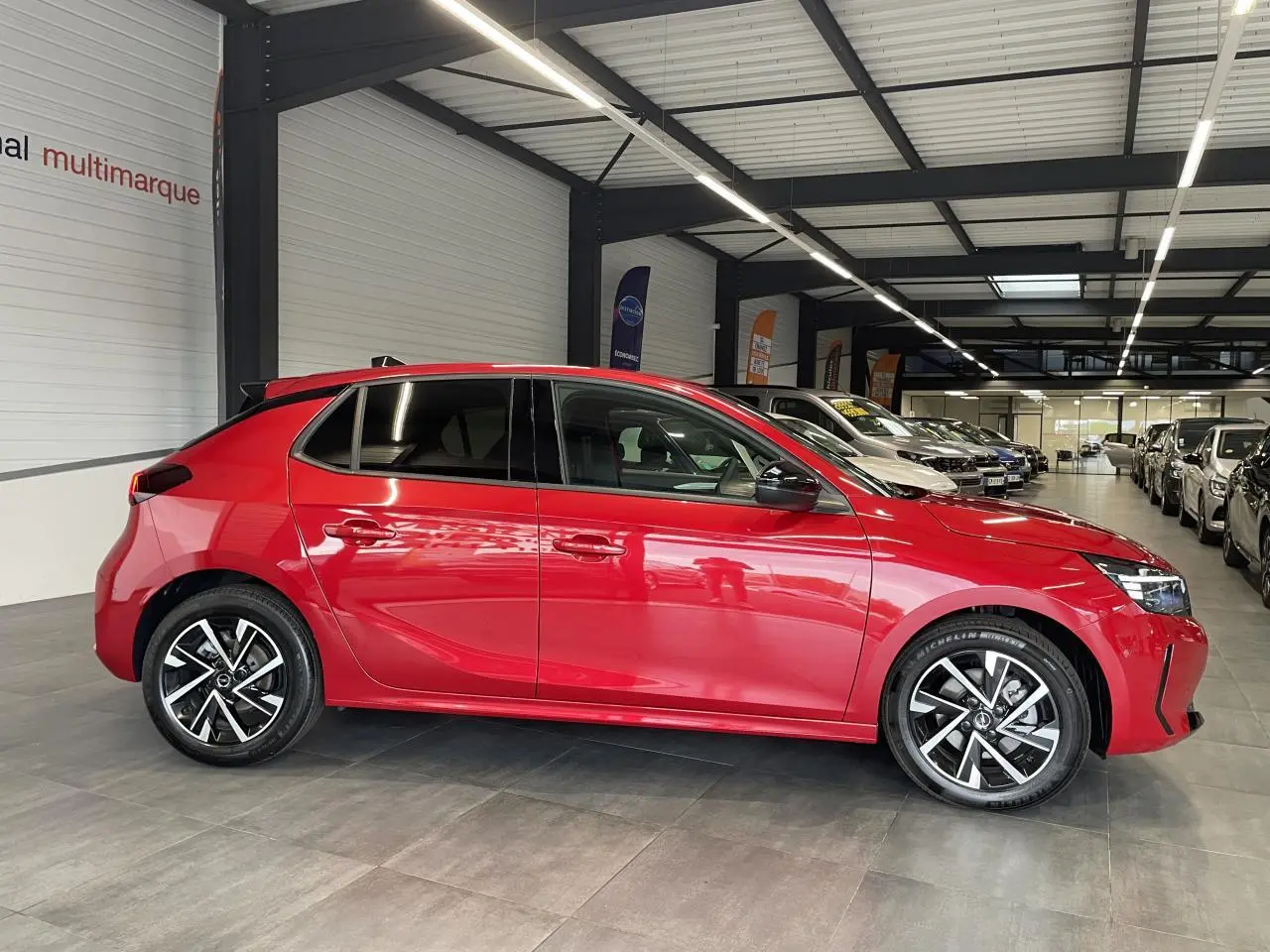 Vue de profil côté gauche d'une Opel Corsa rouge métallisée avec jantes alliage 16 pouces et vitres surteintées en intérieur showroom.