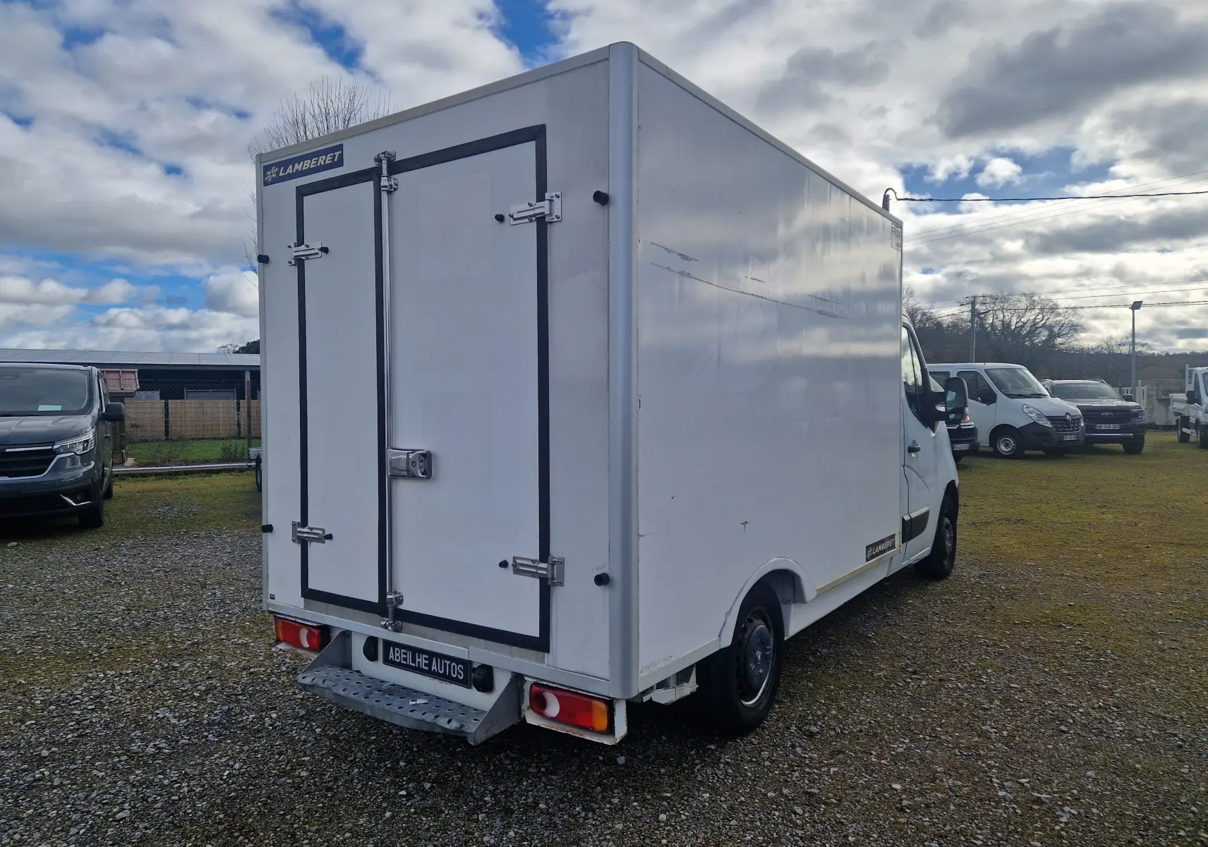 Vue 3/4 arrière droite d'un Renault Master blanc avec cellule frigorifique Lamberet et marchepied arrière sur terrain extérieur.