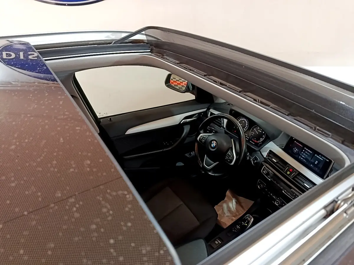 Vue intérieure du poste de conduite d'une BMW X1 noire 2020, avec volant cuir et écran tactile visible depuis le toit ouvrant.