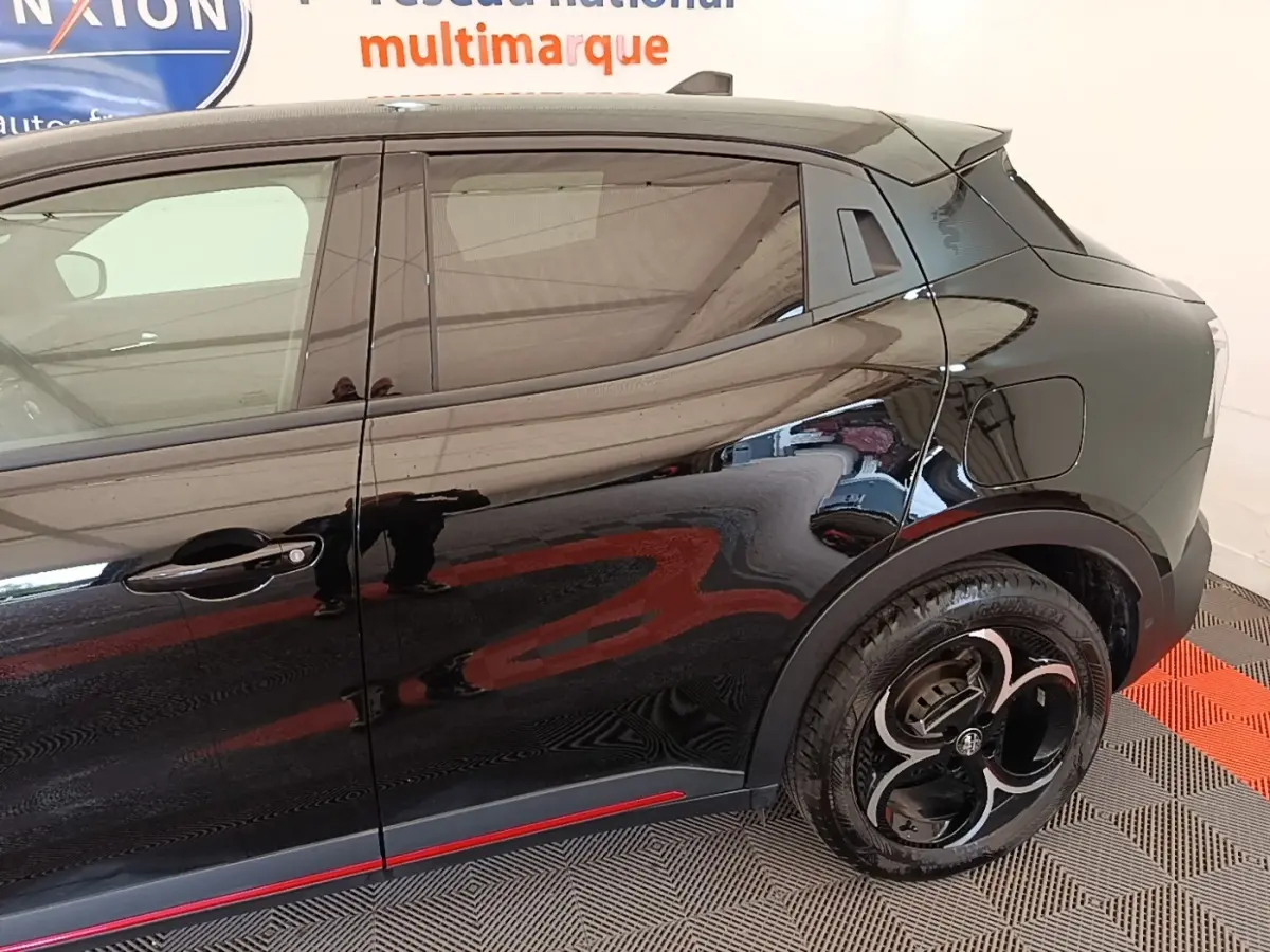 Vue latérale droite de l'Alfa Romeo Junior 1.2 Ibrida noire, avec jantes distinctives et ligne rouge décorative sur le bas de caisse.
