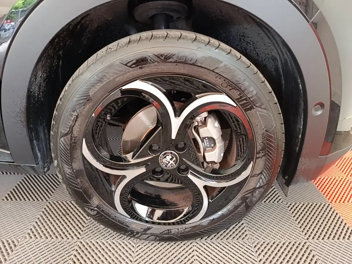 Gros plan sur la roue avant droite noire d'une Alfa Romeo Junior 1.2 Ibrida avec jante au design distinctif en forme de trèfle.