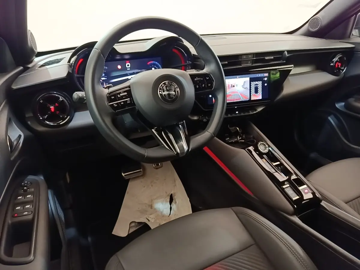Vue intérieure avant gauche de l'Alfa Romeo Junior 1.2 Ibrida 2024, volant cuir, écran tactile et tableau de bord numérique.