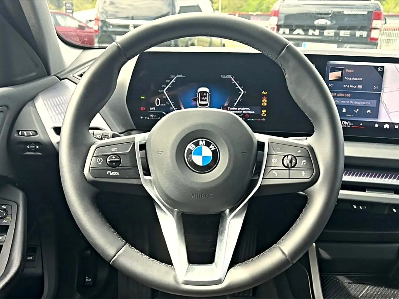 Vue intérieure centrée sur le volant cuir noir BMW d'une Série 1 120da 2025 avec tableau de bord numérique et écran tactile.