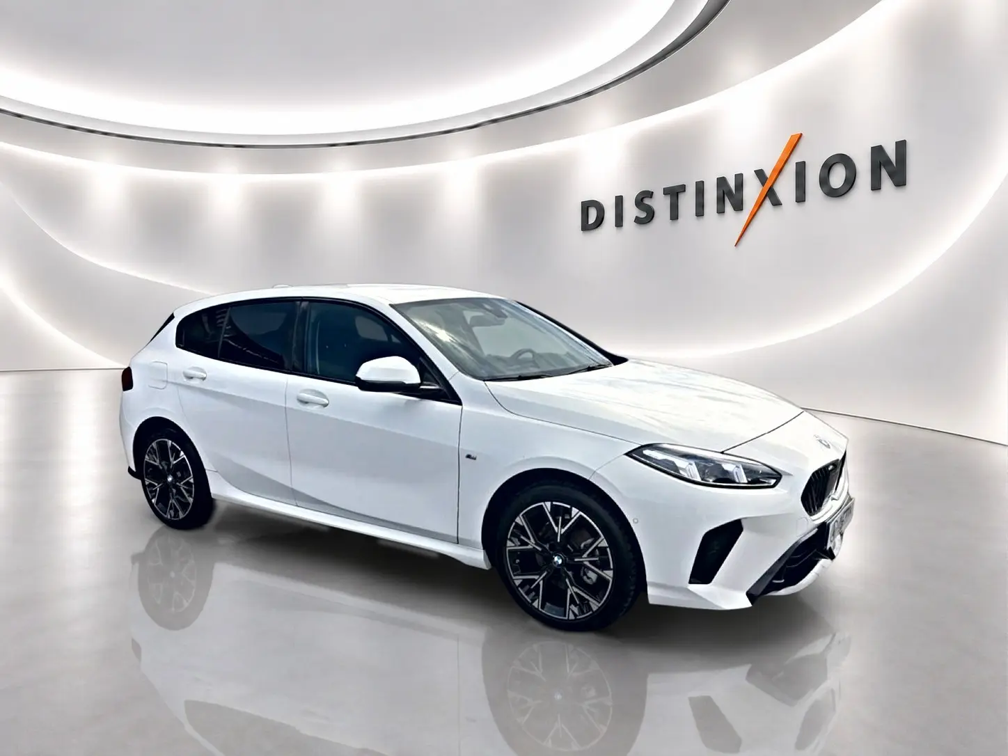BMW Série 1 blanc pur vue 3/4 avant droit dans un showroom lumineux avec jantes noires et logo Distinxion en arrière-plan.