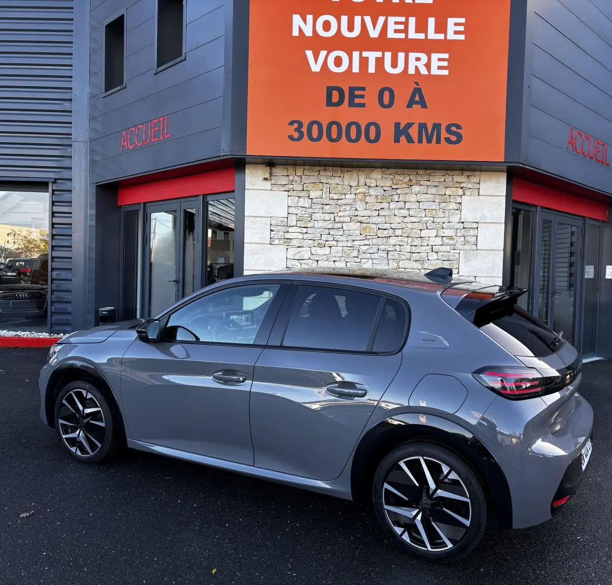 Peugeot 208 Hybrid 136 eDCS6 GT gris foncé vue de profil côté gauche devant un bâtiment avec panneau orange.