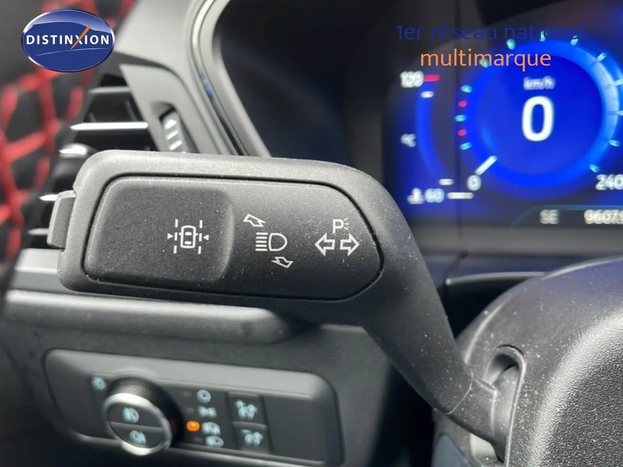 Gros plan sur la manette de clignotant noire à gauche du volant, tableau de bord numérique bleu en arrière-plan.