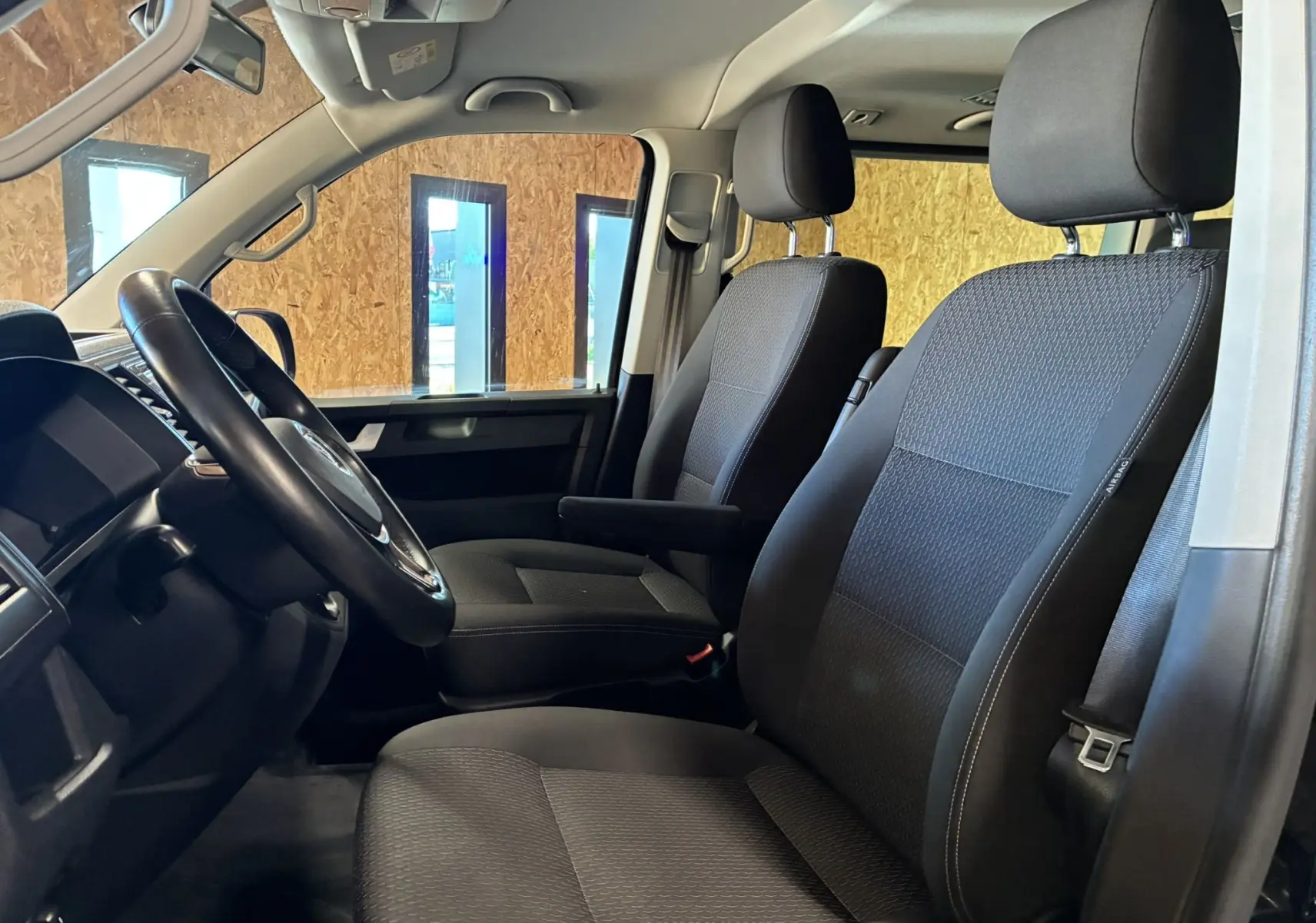 Vue intérieure côté conducteur du Volkswagen Multivan T6 noir, mettant en valeur les sièges avant en tissu noir et le volant.