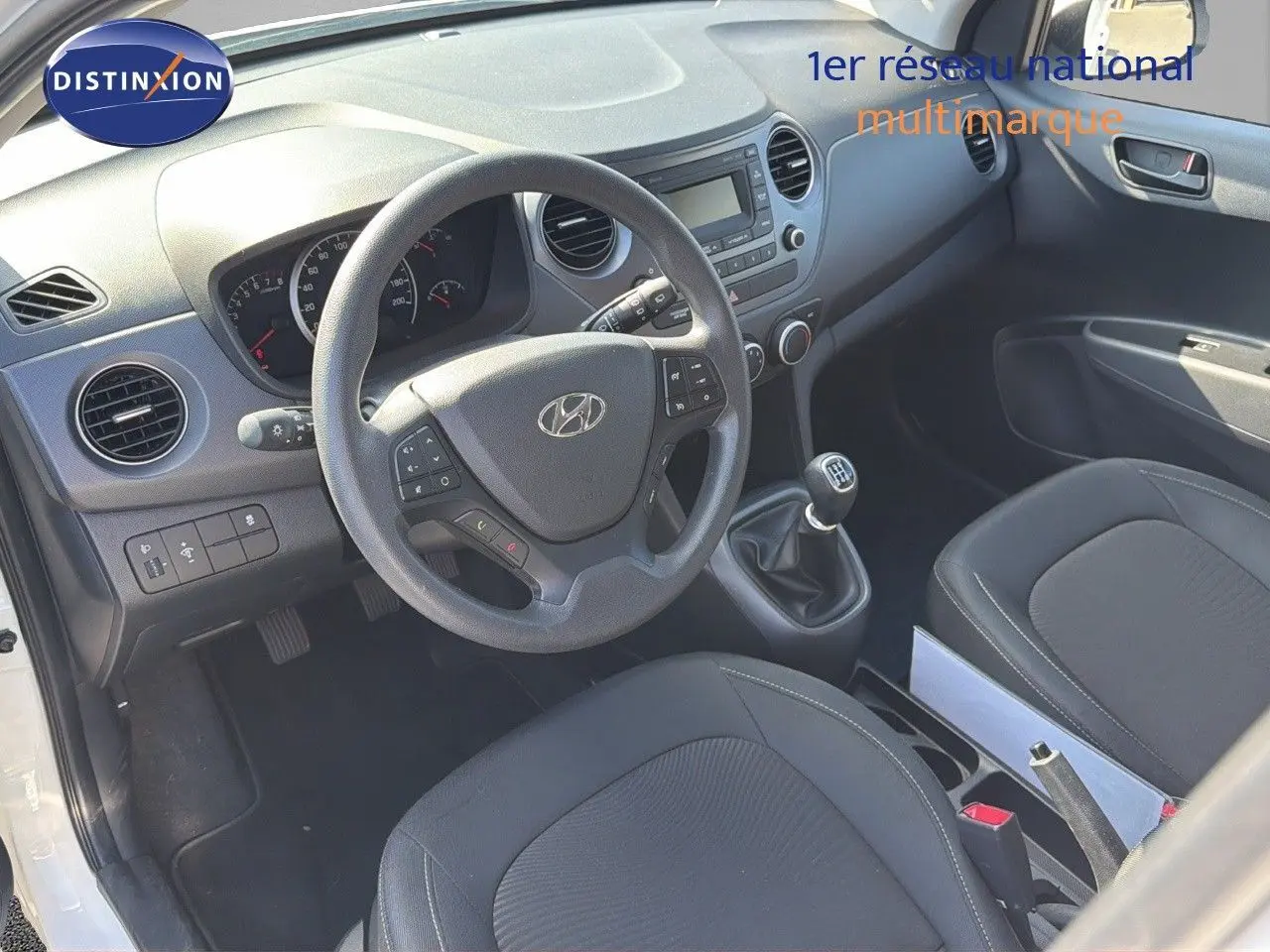 Vue intérieure côté conducteur de la Hyundai i10 blanche 2018, volant multifonction et levier de vitesses manuel visibles.