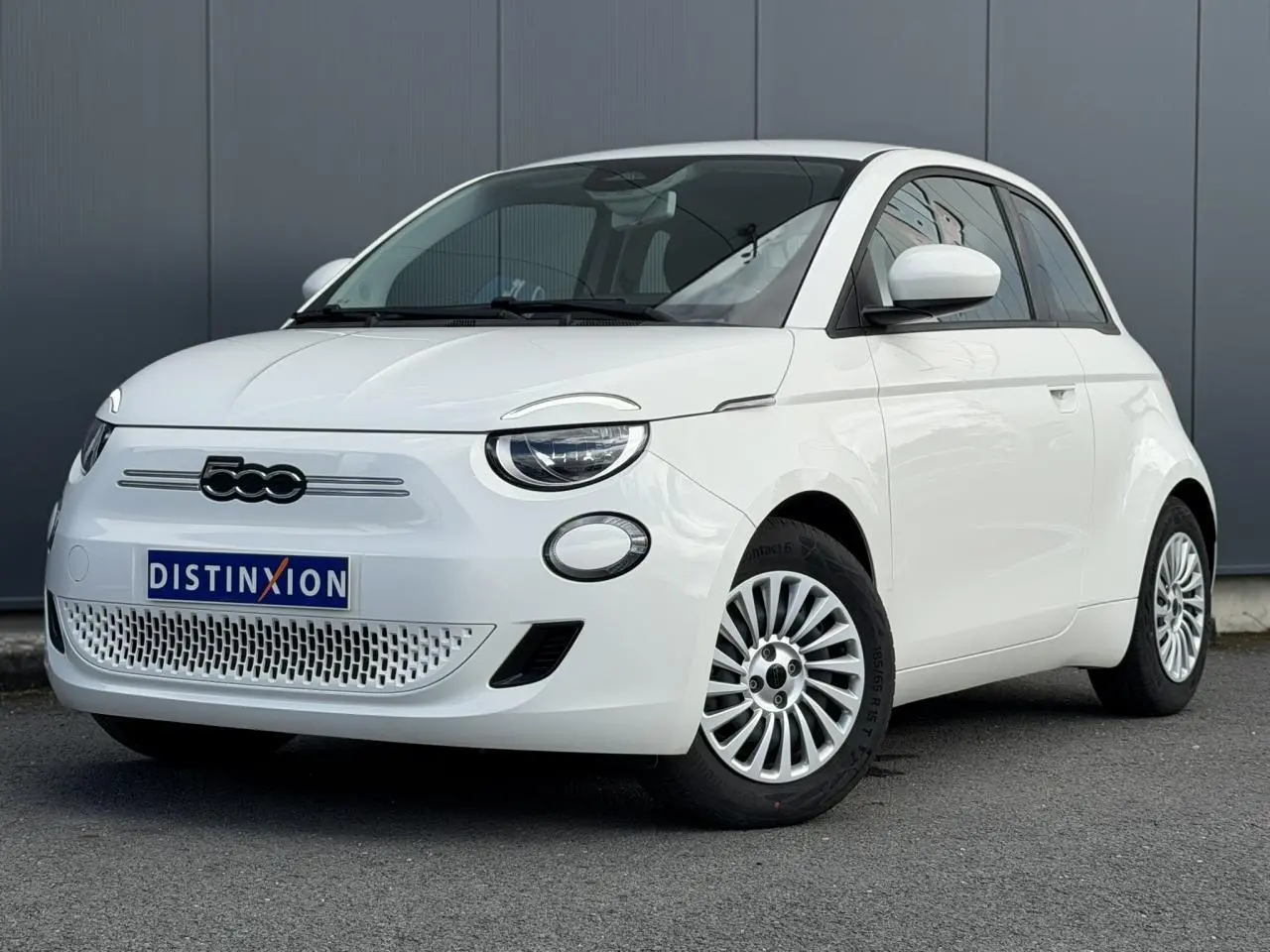 Vue 3/4 avant droite d'une Fiat 500 électrique blanc artic avec jantes alu 16 pouces sur fond urbain.