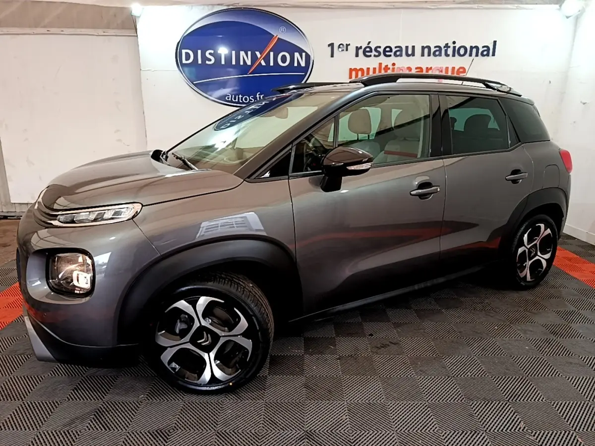 Vue côté gauche 3/4 avant d'un Citroën C3 Aircross gris métallisé avec jantes distinctives et barres de toit noires.