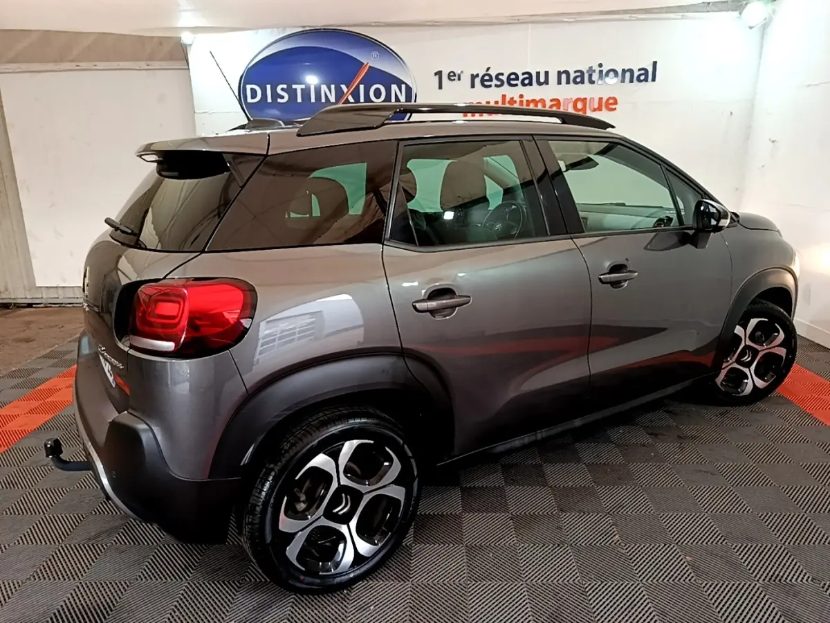 Vue 3/4 arrière droite d'un Citroën C3 Aircross gris métallisé avec barres de toit noires en intérieur showroom.