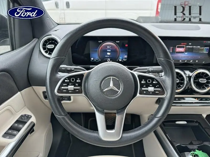 Vue intérieure centrée sur le volant cuir noir de la Mercedes Classe B 250 e avec tableau de bord numérique et garniture beige.