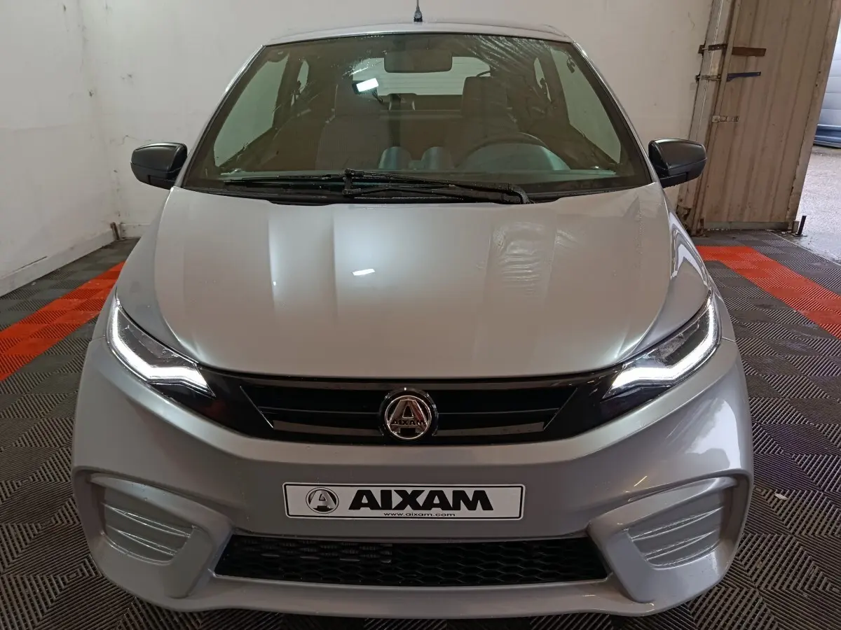 Vue avant d'une Aixam Ambition 0.5 City Pack gris clair avec phares allumés et rétroviseur caméra côté gauche.