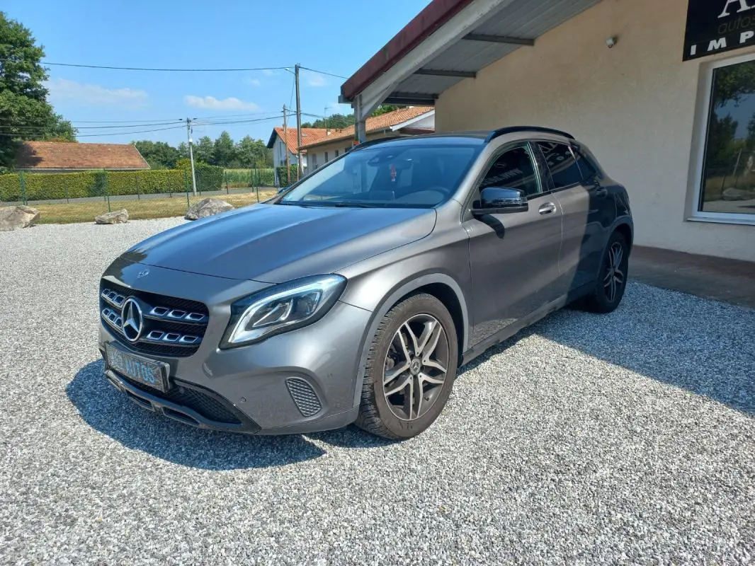 Mercedes GLA gris en 3/4 avant droit, avec calandre distinctive et jantes alliage, stationné sur gravier devant un bâtiment.